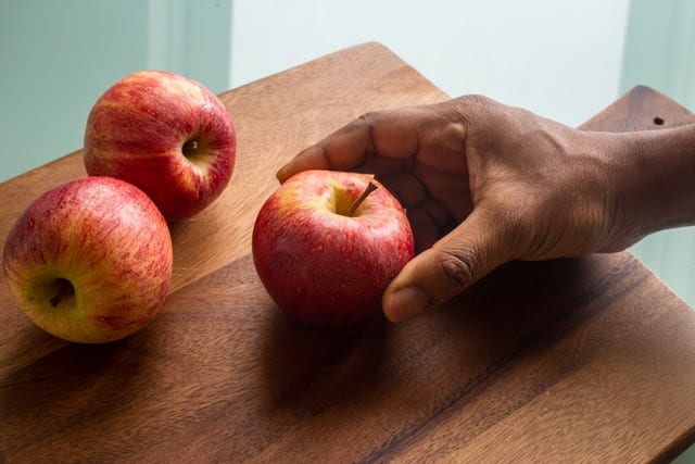 Dłoń, kładąca na drewnianej desce jabłko. Na desce znajdują się już dwa jabłka. 