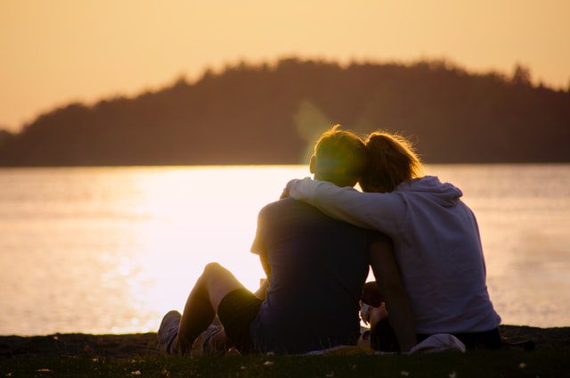 Kobieta przytulona do mężczyzny siedząca nad brzegiem jeziora.