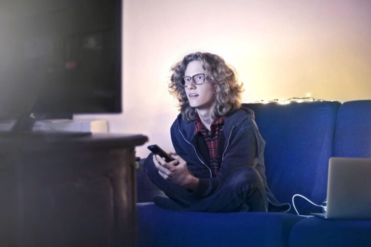 Młody mężczyzna na kanapie ogląda telewizję z telefonem w dłoni